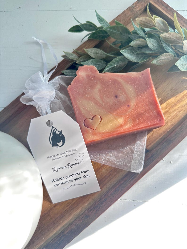 May include: A rectangular bar of handmade goat milk soap with a marbled pink and orange hue, featuring an embossed heart. A white tag attached with a ribbon displays the text: Handmade Goat Milk Soap, TheYummySoap.com, Moroccan Romance.
