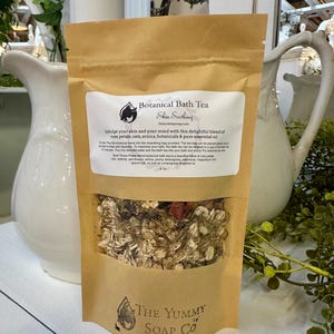 May include: A brown paper pouch of Botanical Bath Tea with a clear window showing dried rose petals, oats, and botanicals. The label features the text Botanical Bath Tea, Skin Soothing, and The Yummy Soap Co, along with usage instructions and ingredients.