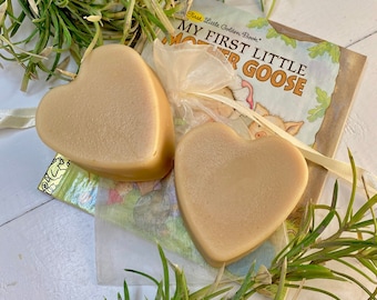 Może przedstawiać: Dwie gładkie, brązowe kostki mydła w kształcie serca umieszczone są na książce First Little Golden Book zatytułowanej My First Little Mother Goose. Pomiędzy mydłami znajduje się przezroczysty biały woreczek z organzy, otoczony zielonymi gałązkami.