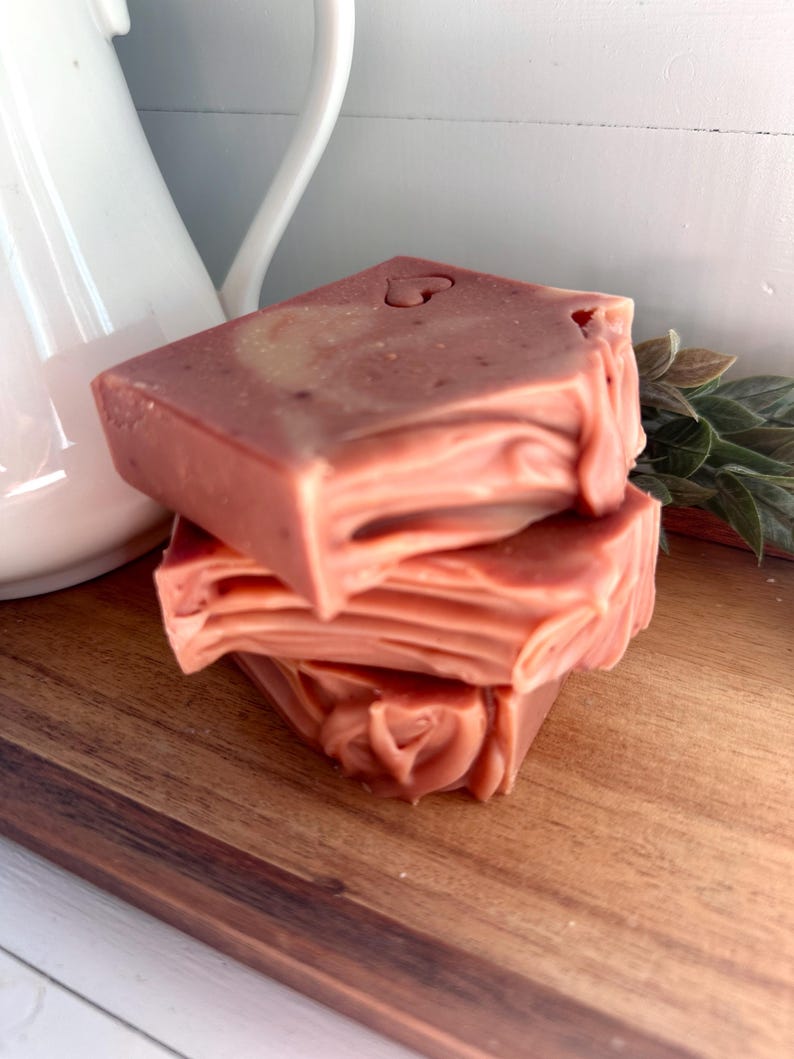 May include: A stack of three rectangular bars of soap in a muted dusty rose color. The soap features a smooth top surface with a small heart-shaped indentation and layered, wavy textures along the sides of each individual bar.