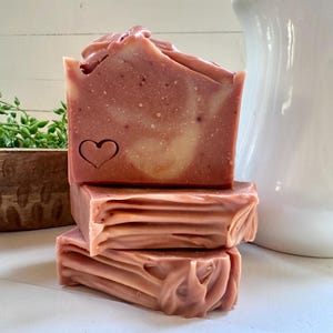 May include: A stack of three rectangular bars of dusty rose soap. The top bar features a stamped heart outline on its front face. The soap has a smooth, marbled texture with subtle swirls of lighter pink and cream throughout the solid block.
