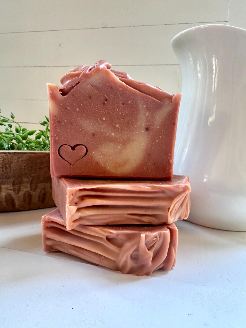 May include: A stack of three rectangular bars of dusty rose soap. The top bar features a stamped heart outline on its front face. The soap has a smooth, marbled texture with subtle swirls of lighter pink and cream throughout the solid block.