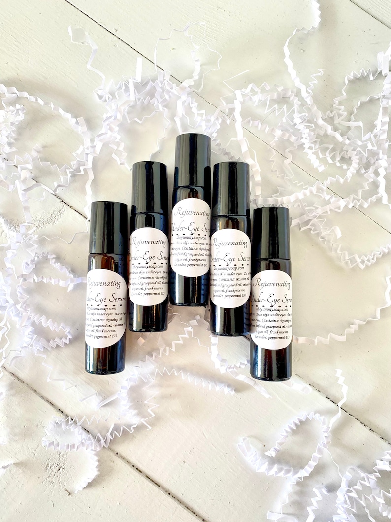 Puede incluir: Cinco peque&ntilde;os frascos de vidrio marr&oacute;n oscuro con bolas rodantes. Cada frasco tiene una etiqueta blanca con el texto "Rejuvenating Under-Eye Serum" y una lista de ingredientes. Los frascos est&aacute;n dispuestos en c&iacute;rculo sobre una superficie blanca.