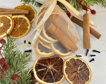 May include: A white organza bag filled with dried orange slices and cinnamon sticks. The bag is tied with a white ribbon and surrounded by additional orange slices, cinnamon sticks, and cloves on a white surface.