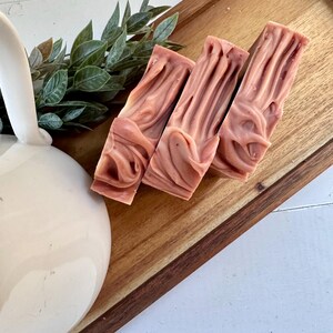 May include: Three rectangular bars of dusty rose soap with a swirled, textured surface. The soap bars are arranged in a row on a wooden tray next to a white ceramic pitcher and a small sprig of green leaves.
