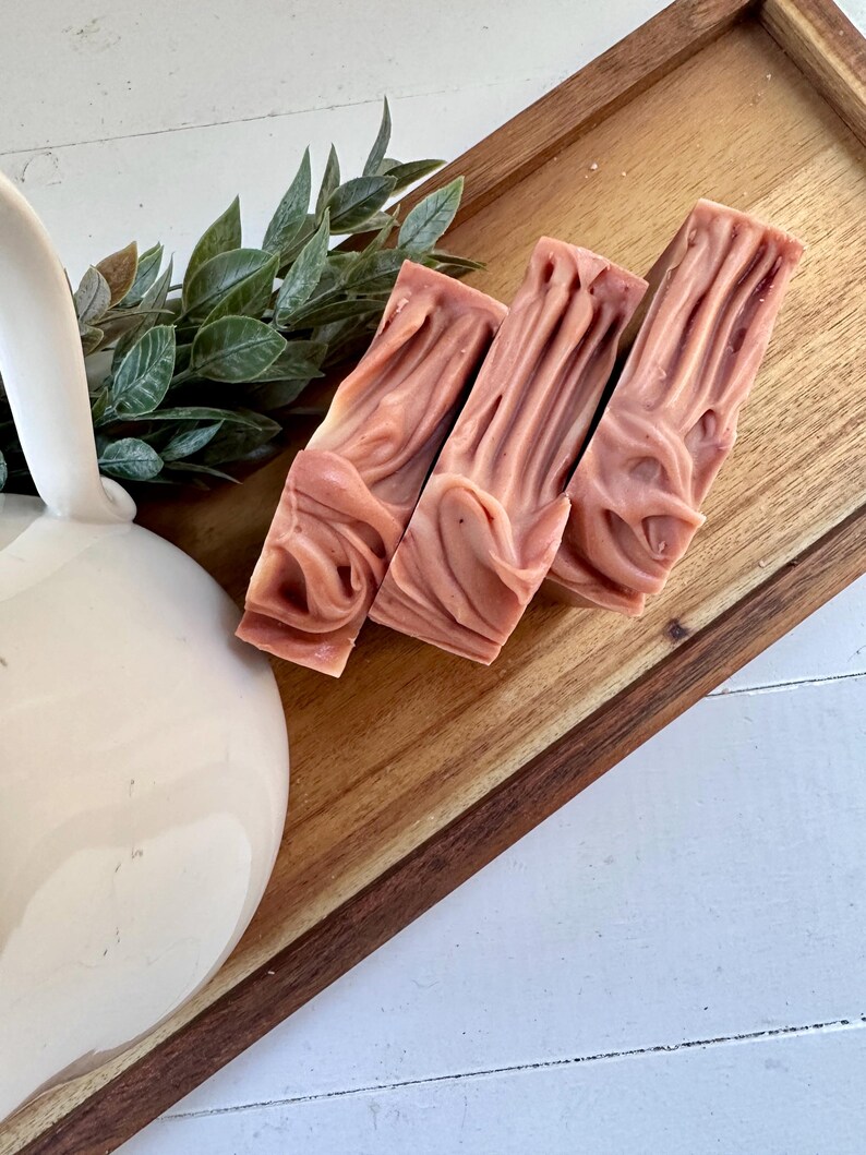 May include: Three rectangular bars of dusty rose soap with a swirled, textured surface. The soap bars are arranged in a row on a wooden tray next to a white ceramic pitcher and a small sprig of green leaves.