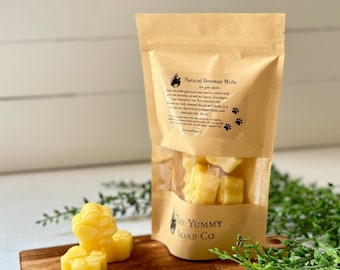 Może przedstawiać: Brązowa papierowa torebka zawierająca żółte woski zapachowe w kształcie łapek. Etykieta zawiera napis Natural Beeswax Melts for pet odors, The Yummy Soap Co. Dwa dodatkowe woski w kształcie łapek znajdują się przed torebką. Dolna część torebki jest przezroczysta.