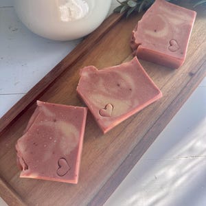 May include: Three rectangular bars of dusty rose soap with a marbled texture and a small heart-shaped indentation on the surface of each piece. The soap bars are arranged in a diagonal line on a rectangular wooden tray with a smooth finish.