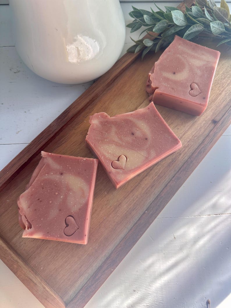 May include: Three rectangular bars of dusty rose soap with a marbled texture and a small heart-shaped indentation on the surface of each piece. The soap bars are arranged in a diagonal line on a rectangular wooden tray with a smooth finish.