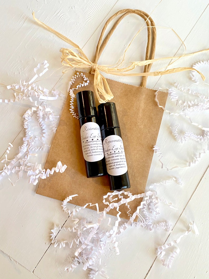 Puede incluir: Dos peque&ntilde;os frascos de vidrio marr&oacute;n con etiquetas negras y texto blanco. Los frascos est&aacute;n etiquetados como "Restoring Under-Eye Serum" y "Rejuvenating Under-Eye Serum". Los frascos est&aacute;n en una bolsa de papel marr&oacute;n con una manija de papel marr&oacute;n y un lazo de papel marr&oacute;n. La bolsa est&aacute; rodeada de trozos de papel blanco.
