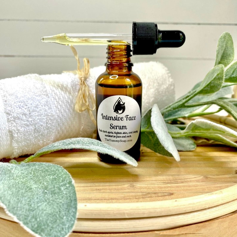 May include: A brown glass bottle of Intensive Face Serum with a black dropper. The label reads "Intensive Face Serum" and "TheYummySoap.com". A white towel and green leaves are in the background. The serum is designed to fade dark spots, lighten skin, and moisturize the face and neck.