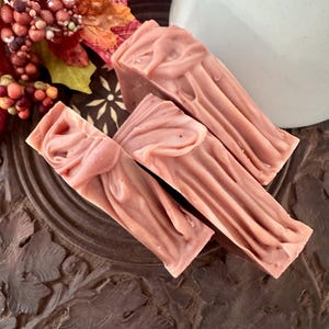 May include: Three rectangular bars of dusty rose-colored soap with a swirled, textured top surface. The soap bars are arranged in a cluster, showcasing a smooth, matte finish and a consistent muted pink hue throughout each individual piece.