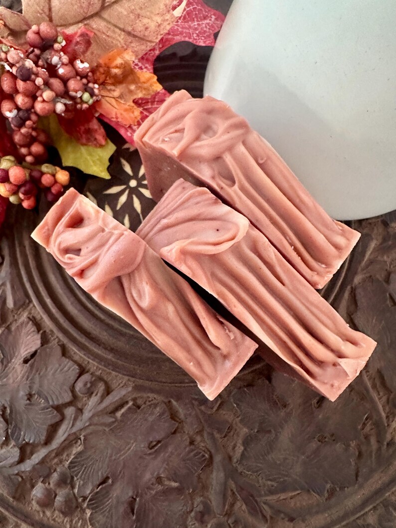 May include: Three rectangular bars of dusty rose-colored soap with a swirled, textured top surface. The soap bars are arranged in a cluster, showcasing a smooth, matte finish and a consistent muted pink hue throughout each individual piece.