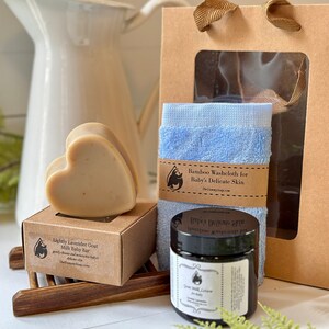 Może przedstawiać: Zestaw upominkowy zawierający mydło w kształcie serca Lightly Lavender Goat Milk Baby Bar w brązowym pudełku, niebieską myjkę bambusową z papierową opaską Bamboo Washcloth for Baby's Delicate Skin oraz słoiczek balsamu dla niemowląt.