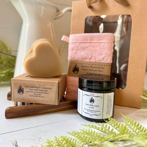Może przedstawiać: Zestaw upominkowy zawierający mydło z koziego mleka w kształcie serca w brązowym pudełku z etykietą Pure and Simply Unscented Goat Milk Baby Bar, r&oacute;żową myjkę bambusową, słoiczek balsamu z koziego mleka oraz brązową papierową torebkę prezentową.