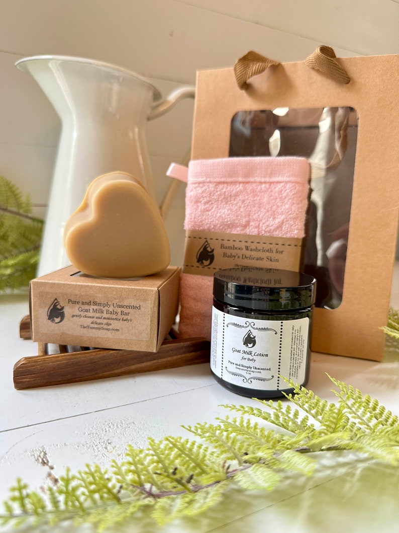Może przedstawiać: Zestaw upominkowy zawierający mydło z koziego mleka w kształcie serca w brązowym pudełku z etykietą Pure and Simply Unscented Goat Milk Baby Bar, r&oacute;żową myjkę bambusową, słoiczek balsamu z koziego mleka oraz brązową papierową torebkę prezentową.
