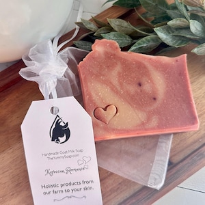May include: A rectangular bar of pink and cream marbled goat milk soap with a small heart-shaped indentation. Attached is a white tag with a black goat logo, the text "Handmade Goat Milk Soap", "Moroccan Romance", and "Holistic products from our farm to your skin."