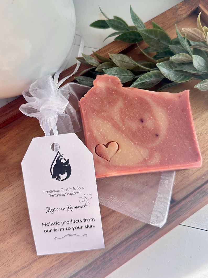 May include: A rectangular bar of pink and cream marbled goat milk soap with a small heart-shaped indentation. Attached is a white tag with a black goat logo, the text "Handmade Goat Milk Soap", "Moroccan Romance", and "Holistic products from our farm to your skin."
