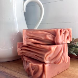 May include: A stack of three rectangular bars of dusty rose colored soap with a marbled, swirled texture. The soap bars are positioned in front of a white ceramic pitcher and a small green plant on a wooden surface.