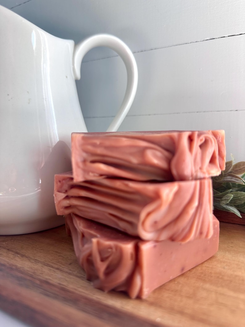 May include: A stack of three rectangular bars of dusty rose colored soap with a marbled, swirled texture. The soap bars are positioned in front of a white ceramic pitcher and a small green plant on a wooden surface.