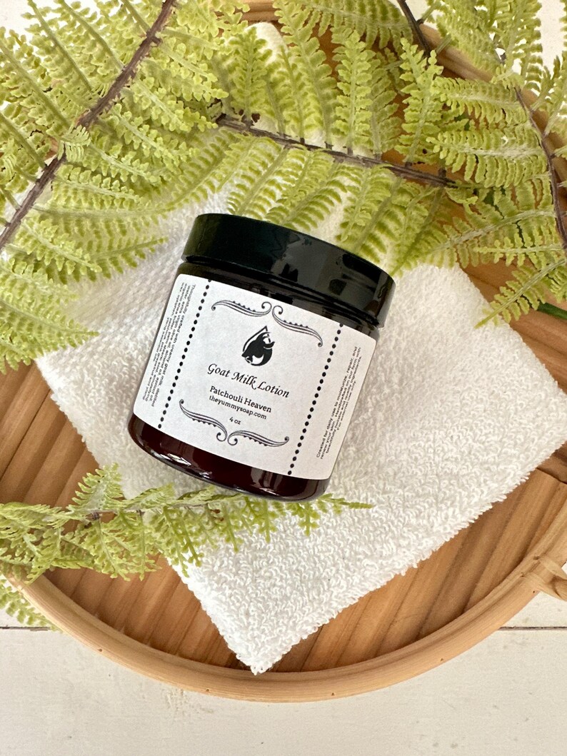May include: A dark amber glass jar of Goat Milk Lotion with a black screw-top lid. The white label features a black goat icon, the text Goat Milk Lotion, Patchouli Heaven, theyummysoap.com, and 4 oz, framed by decorative black scrollwork.