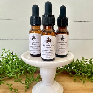 May include: Three amber glass bottles of Nail and Cuticle Serum with black droppers are displayed on a white pedestal. Each bottle has a label with product information and a logo. The bottles are surrounded by greenery.