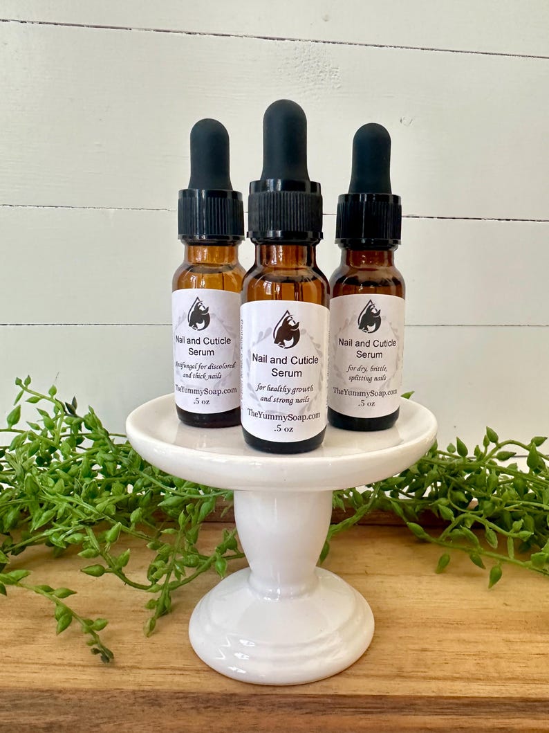May include: Three amber glass bottles of Nail and Cuticle Serum with black droppers are displayed on a white pedestal. Each bottle has a label with product information and a logo. The bottles are surrounded by greenery.