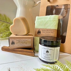 Może przedstawiać: Beżowe mydło w kształcie serca na kartonowym pudełku z etykietą Freshly Tea Tree Goat Milk Baby Bar, jasnozielona myjka bambusowa oraz słoiczek Goat Milk Lotion for Baby, zaprezentowane z brązową papierową torebką prezentową i drewnianą podstawką.