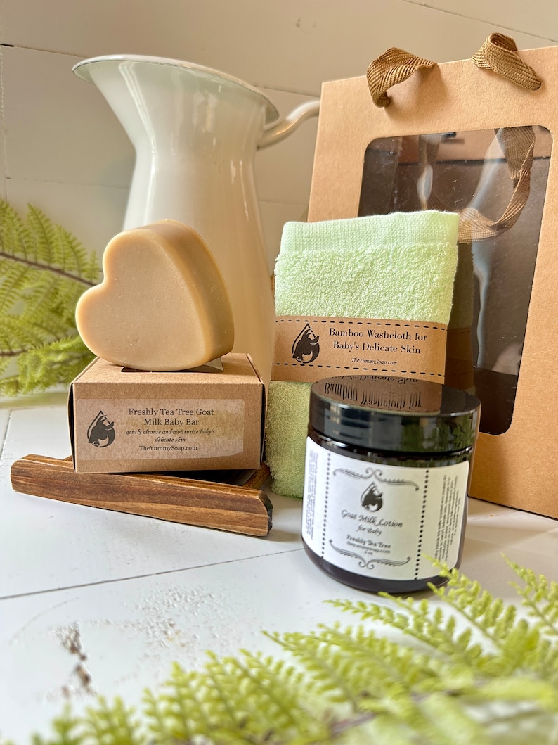 Może przedstawiać: Beżowe mydło w kształcie serca na kartonowym pudełku z etykietą Freshly Tea Tree Goat Milk Baby Bar, jasnozielona myjka bambusowa oraz słoiczek Goat Milk Lotion for Baby, zaprezentowane z brązową papierową torebką prezentową i drewnianą podstawką.