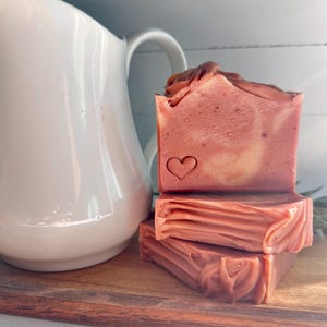 May include: A stack of three rectangular bars of dusty rose-colored soap with a smooth texture and a small heart-shaped stamp on the front face. The soap bars are positioned next to a white ceramic pitcher with a curved handle.
