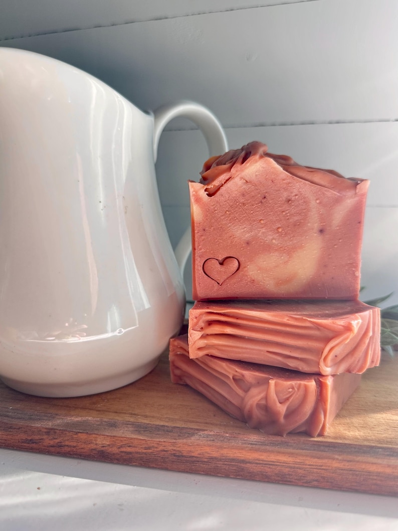 May include: A stack of three rectangular bars of dusty rose-colored soap with a smooth texture and a small heart-shaped stamp on the front face. The soap bars are positioned next to a white ceramic pitcher with a curved handle.