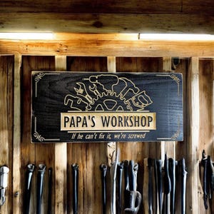 May include: A black wooden sign with gold lettering that reads "Papa's Workshop" and "If he can't fix it, we're screwed". The sign is decorated with a gold silhouette of tools and a gear.