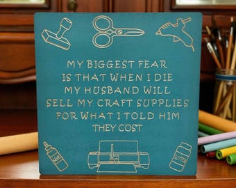 May include: A square teal wooden sign featuring engraved icons of scissors, a glue gun, a glue bottle, a cutting machine, and a glitter jar. The text reads: MY BIGGEST FEAR IS THAT WHEN I DIE MY HUSBAND WILL SELL MY CRAFT SUPPLIES FOR WHAT I TOLD HIM THEY COST.