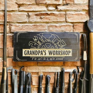 May include: A wooden sign with the words "GRANDPA'S WORKSHOP" and the phrase "If he can't fix it, we're screwed." The sign features a gold-colored border and a graphic of tools. The sign is mounted on a brick wall.