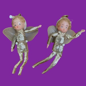 May include: Two identical elf figurines with blonde hair, wearing metallic gold suits with white pom-pom accents. Each elf features translucent wings, a pointed hat, and a painted face with blue eyes and an open mouth, posed with arms outstretched.