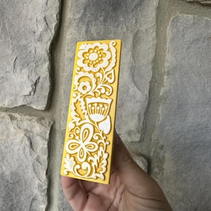 May include: A rectangular, yellow decorative object with a white floral design. The object features a raised, intricate pattern of flowers, leaves, and swirling lines. The object is held in a hand against a stone background.