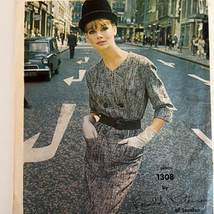 May include: A vintage Vogue Couturier Design pattern for a dress, pattern number 1308 by Ronald Paterson of London. The pattern features a black and white checkered dress with a black belt and white gloves. The model is wearing a black hat and is standing on a street.
