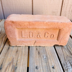 May include: A rectangular terracotta-colored clay brick featuring a recessed central panel with the embossed text L.D. & Co. The brick has a rough, porous texture and weathered edges, displaying a natural reddish-brown hue throughout its surface.