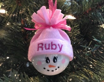 May include: A white snowman ornament with a pink hat and a pom-pom. The hat has the name "Ruby" embroidered on it.