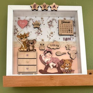 Puede incluir: Una caja de sombra con marco blanco, con recortes de madera de una jirafa, un koala y una niña en un balancín. Incluye un calendario, hora, peso (3.300 kg) y longitud (51 cm). Se muestra el nombre "Eva".