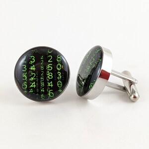 May include: A pair of silver cufflinks with black and green circular inlays. The inlays feature a pattern of green numbers on a black background.
