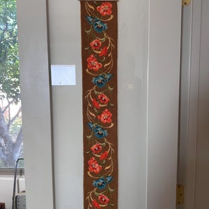 May include: A long, rectangular tapestry with a brown background and a floral design. The flowers are red and blue, with green stems and leaves. The tapestry is hanging on a white door.