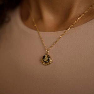 Pode incluir: Um colar de corrente de ouro com um pingente preto e dourado com um design detalhado. O colar é usado por uma pessoa com uma blusa marrom claro.