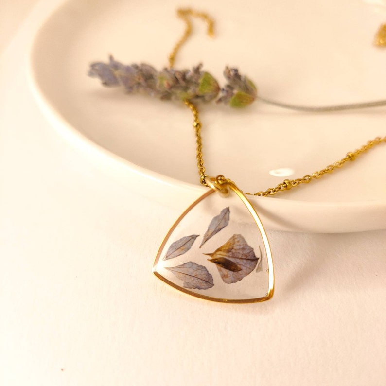 May include: A gold chain necklace with a triangular pendant featuring pressed purple flowers encased in clear resin.