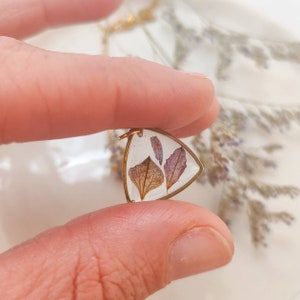 Könnte beinhalten: Ein goldener Dreiecksanhänger mit gepressten Trockenblumen. Die Blumen sind eine Mischung aus Braun und Lila.