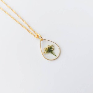 May include: A gold chain necklace with a teardrop-shaped pendant featuring a pressed white flower inside.