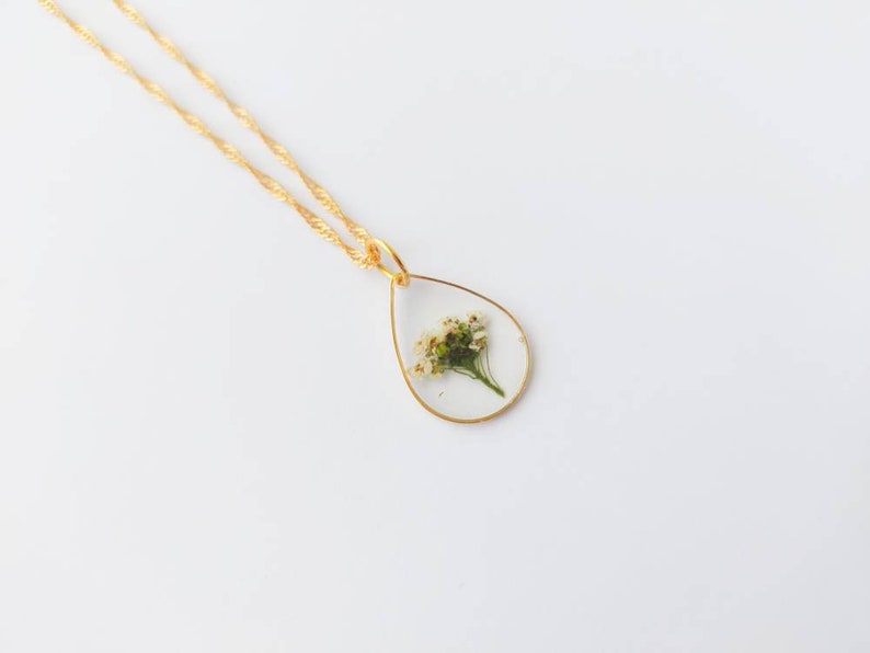 May include: A gold chain necklace with a teardrop-shaped pendant featuring a pressed white flower inside.