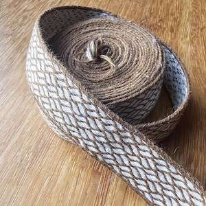 May include: A roll of decorative ribbon with a woven pattern. The ribbon features a brown and white diamond design, with a brown border. The ribbon is rolled up on a wooden surface.