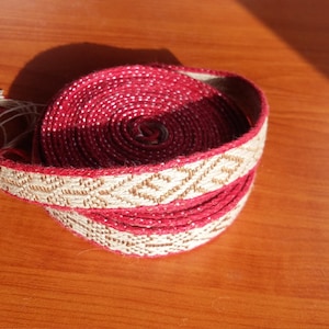 May include: A red and white woven ribbon with a geometric pattern. The ribbon is rolled up and tied with a white tassel.