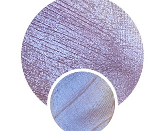 May include: A close-up of a light purple eyeshadow with a shimmery finish. The eyeshadow is in a round pan and has a textured surface.